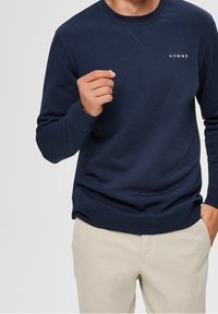 Námořnická modrá mikina s kulatým výstřihem, dlouhými rukávy a žebrovanými manžetami. Má malé bílé logo "HOMME" na hrudi. Materiál z bavlněné směsi.