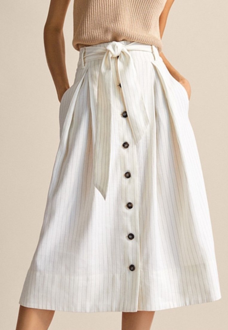 Femme portant une jupe midi blanche boutonnée sur le devant, avec de fines rayures verticales et une ceinture en tissu nouée à la taille.
