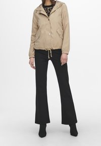 Woman wearing beige hooded jacket with front pockets, black flared pants, and black textured ankle boots standing against white background.