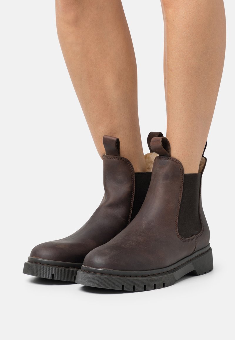 Botas Chelsea de cuero marrón con paneles laterales elásticos, una suela de goma resistente y lengüetas para facilitar su puesta. La superficie tiene una textura suave.
