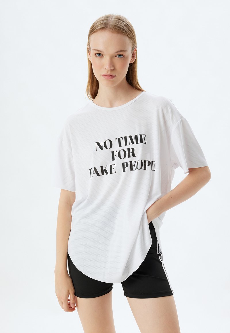 Hvid oversized kortærmet t-shirt med sort tekst "NO TIME FOR FAKE PEOPLE"; tekstureret stof med en rund nederkant, parret med sorte shorts.