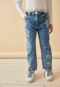 Jeans de mezclilla azul claro con grandes parches de estrellas brillantes, de cintura alta, corte recto, con dobladillo desflecado, combinados con zapatillas blancas y un top con detalles de encaje.