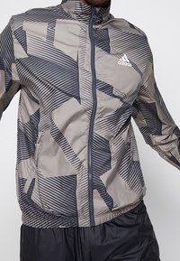 Jasnoszara kurtka przeciwwiatrowa z czarnymi geometrycznymi wzorami, z pełnym zamkiem, wysokim kołnierzem, elastycznymi mankietami i dołem. Z logo Adidas.