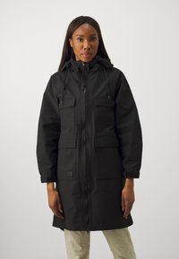 Manteau noir à capuche avec fermeture éclair, deux poches sur la poitrine et poignets réglables. Texture lisse avec une coupe décontractée et un design long jusqu'au genou.