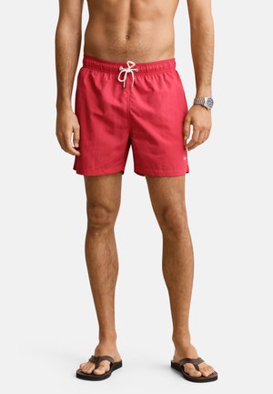 Uomo che indossa pantaloncini da bagno rossi con cordoncino bianco, infradito marroni e orologio da polso argento, in piedi davanti a uno sfondo semplice.