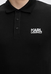 Μαύρο πόλο πουκάμισο με τρία κουμπιά και λευκό λογότυπο "KARL LAGERFELD" στην αριστερή πλευρά του στήθους.
