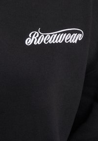 Crna dukserica s rebrastom teksturom, sa bijelim izvezenim "Rocawear" logom sprijeda. Jednostavan, klasičan dizajn.
