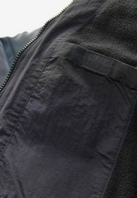 Schwarze Jacke mit einem glatten, glänzenden Außenmaterial und einem strukturierten Fleece-Panel. Enthält einen Reißverschluss und eine Taschenleiste.