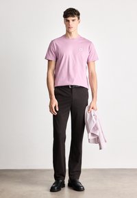 Lys rosa bomull t-skjorte med en liten logo, sammen med mørkebrune bukser og svarte sko. Holder en lysstripete langermet skjorte.