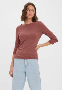 Brązowa bluzka z trzema czwarciami rękawów, wykonana z gładkiego materiału, z dekoltem typu łódka, w połączeniu z jasnoniebieskimi spodniami typu wide-leg.