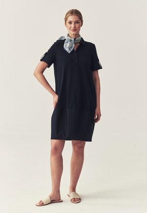 Donna in piedi che indossa un vestito blu navy a maniche corte, una sciarpa da collo a fantasia blu e bianca e sandali beige con fascia su sfondo beige.