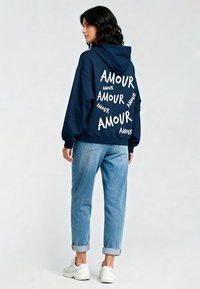 Felpa navy con testo bianco "AMOUR" in varie misure, abbinata a jeans blu chiaro con risvolti e sneakers bianche. Tessuto morbido, vestibilità casual.