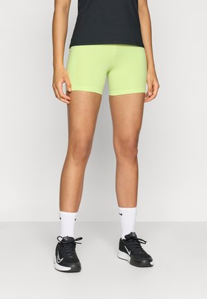 Pantaloncini da atleta verde neon, design aderente, tessuto morbido, abbinati a scarpe da ginnastica nere e calzini bianchi. Senza motivi o accenti evidenti.