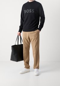 Felpa nera con grande logo grigio "BOSS", pantaloni beige sartoriali e borsa tote in pelle nera. Sneakers bianche completano l'outfit.