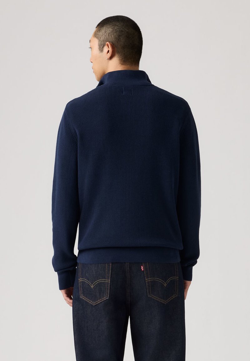 Marineblauer Pullover aus geripptem Strick mit hohem Kragen und langen Ärmeln, kombiniert mit dunklen Jeans aus Denim mit aufgenähten Gesäßtaschen.