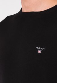 Maglione nero in morbido tessuto a maglia, caratterizzato da un collo rotondo. Logo bianco "GANT" e stemma sulla parte sinistra del petto.