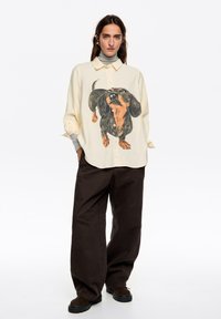 Camisa oversize color crema con una gran ilustración de un dachshund, cierre de botones en la parte delantera, combinada con pantalones oscuros y holgados y calzado marrón.