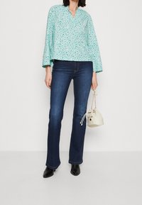 En turkos blommig blus med V-ringning och utsvängda ärmar, tillsammans med mörkblå utsvängda jeans och en krämfärgad bucket bag. Svarta ankelskor avslutar looken.