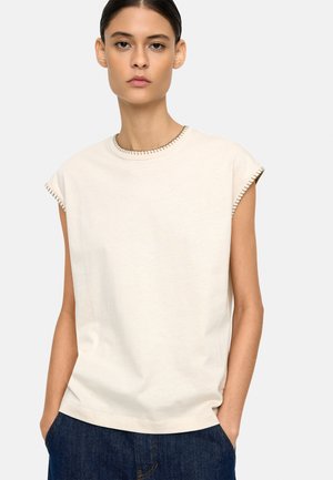 Kobieta o ciemnych włosach, w białej bezrękawnikowej bluzce z przeszywanymi brzegami, z rękami w kieszeniach, nosząca niebieskie dżinsy, zwrócona przodem na jednolitym tle.