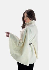 Blusa tipo capa de tejido blanco acanalado con cuello alto y mangas largas, ajustada en la cintura con un cinturón negro. Textura suave y corte relajado.