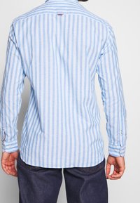 Light blue and white striped long-sleeve shirt with a button-down collar, rounded hem, and two button cuffs. Fabric texture appears smooth.