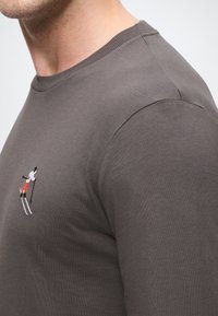 Pilkas ilgomis rankovėmis džemperis su pynimo apykakle ir slidinėjimo tema siuvinėtu raštu raudona, juoda ir balta spalvomis krūtinėje.