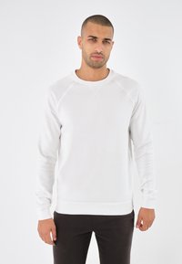 Sudadera blanca con cuello redondo y mangas largas. Presenta costuras de estilo raglán y un acabado texturizado. Combinada con pantalones oscuros ajustados.
