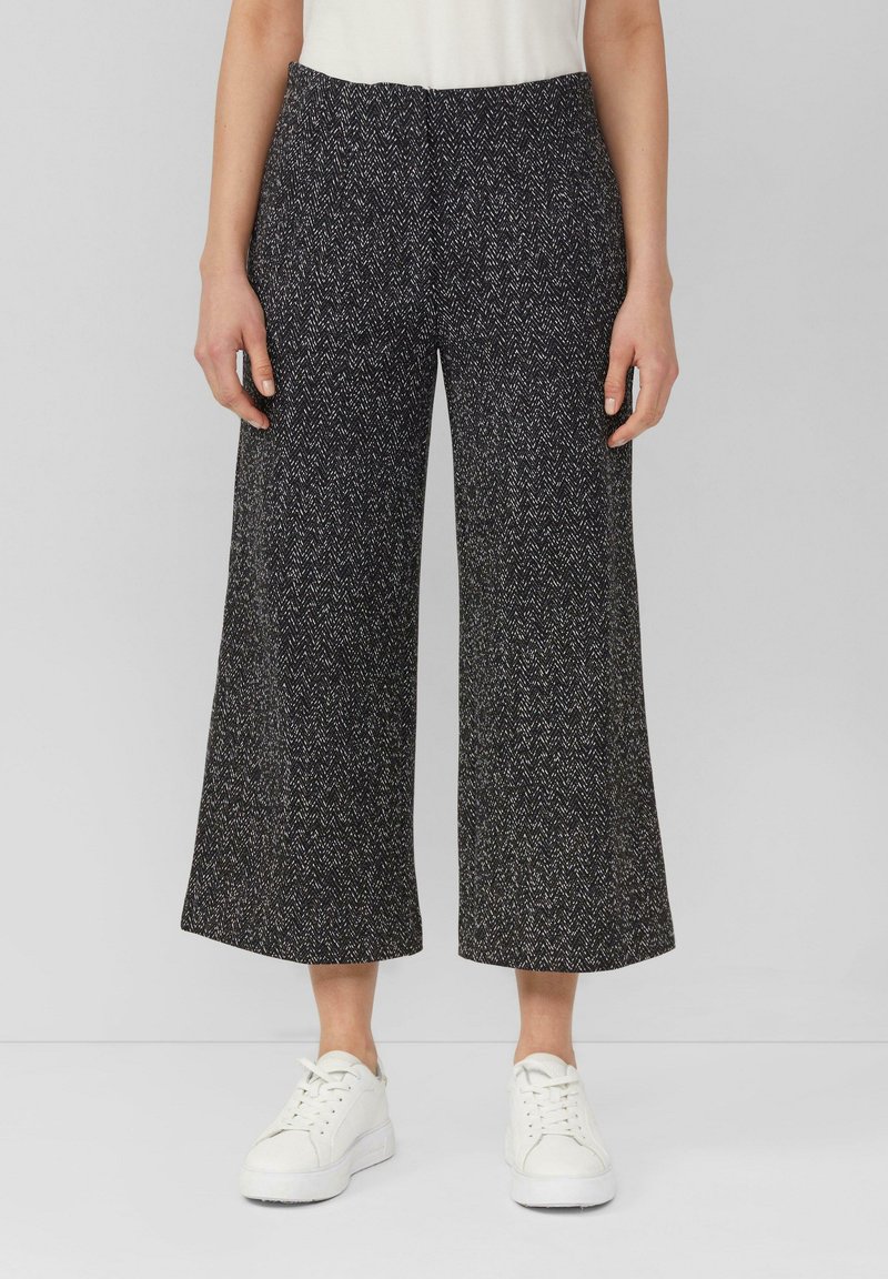 Femme portant un pantalon large court en chevron noir et blanc avec des baskets blanches sur fond uni.