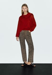 Vrouw staat met haar hand in haar zak, gekleed in een losse rode gebreide trui, bruine suède broek en zwarte open-toe sleehakken op een zwarte vloer.