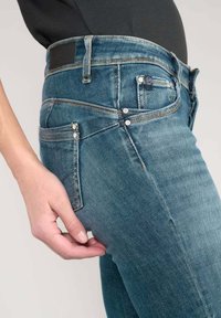 Jean en denim bleu avec une coupe slim, dotés d'une taille mi-haute, de cinq poches et de coutures contrastées. Texture lisse avec un aspect légèrement utilisé.