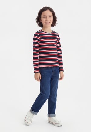 Barn står, smilende, iført en rød, hvit og marineblå stripete langermet skjorte, blå oppbrettede jeans og hvite joggesko.