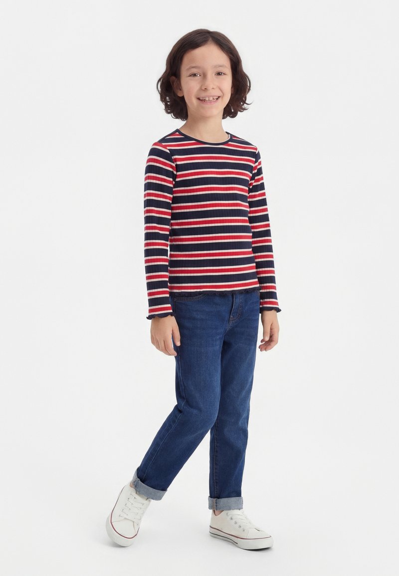 Παιδί που στέκεται, χαμογελώντας, φορώντας μία μακρυμάνικη μπλούζα με κόκκινες, άσπρες και ναυτικές ρίγες, μπλε τζιν με ρεβέρ και λευκά αθλητικά παπούτσια.