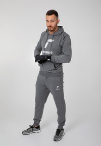 Grå hættetrøje med hvidt logo, parret med matchende grå joggingbukser. Handskerne er sorte. Sneakersne er grå med sorte detaljer. Enkel, atletisk design.