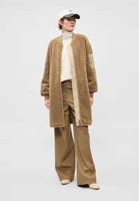 Brūns, pūkains, garš mētelis ar kviltētu priekšpusi, kombinēts ar bēšām platām biksēm un krēmkrāsas augstu apkakli, papildināts ar zaļu un krēmkrāsas cepuri.