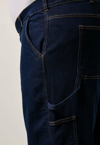 Pantalones de mezclilla azul oscuro con un bolsillo lateral y costuras amarillas en contraste, de corte clásico. Textura suave y colocación estándar de los bolsillos.