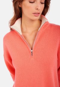 Pull-over mi-zip en corail fabriqué en tissu tricoté texturé, avec un col intérieur blanc contrastant et une fermeture éclair argentée.