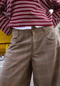 Szerokie spodnie w kratę w odcieniach brązu, beżu i pomarańczu; z zapięciem na guzik i przednimi kieszeniami. Częściowo widoczna paskowana bordowa bluzka.