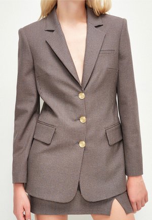 Femme portant un blazer à carreaux marron avec des boutons dorés et une jupe assortie avec une fente sur le devant, debout devant un fond uni.