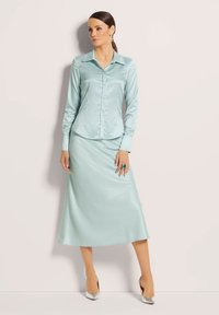 Een lichtblauwe satijnen blouse met een kraag en knoopsluiting aan de voorkant, gecombineerd met een bijpassende satijnen midi-rok en zilveren hakken.