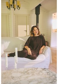 Femme en tenue plissée sombre, allongée sur un canapé blanc dans un salon lumineux et minimaliste avec des bougies et un lustre au plafond.