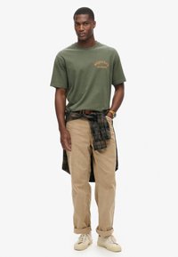 Camiseta de algodón verde oliva con logotipo naranja, combinada con pantalones de pana beige y una camisa de cuadros azul atada a la cintura. Zapatillas cremosas.