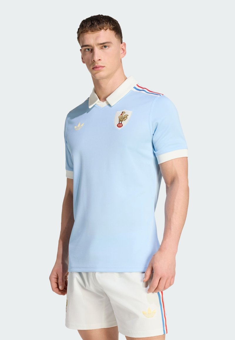 Young man wearing light blue polo shirt with white collar and shorts, featuring red and blue stripes and a rooster emblem.