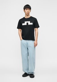 Μαύρο κοντομάνικο T-shirt με λευκό γραφικό σχέδιο στο μπροστινό μέρος, συνδυασμένο με ανοιχτό μπλε, φαρδιά τζιν παντελόνια και μαύρα αθλητικά παπούτσια.