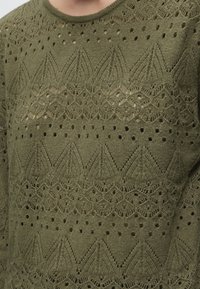 Pull vert en maille avec des motifs ajourés complexes aux designs géométriques et floraux sur la poitrine et les manches.