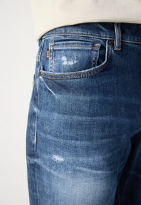 Blue denim jeans with a faded, slightly distressed texture. Features orange stitching and a visible brand tag on the back pocket.