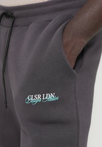 Calças de moletom cinzentas feitas de um tecido macio, apresentando um cós com cordão e bolsos laterais. Inclui texto bordado em branco e turquesa.