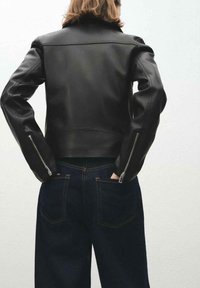 Czarna skórzana kurtka o dopasowanym kroju, z zamykanymi na suwak mankietami i wysokim kołnierzem, w połączeniu z ciemnymi spodniami dżinsowymi. Gładka faktura i dopasowany kształt.