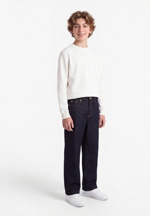 Adolescent aux cheveux bouclés portant une chemise blanche à manches longues, un jean foncé et des baskets blanches, debout et souriant sur fond blanc.