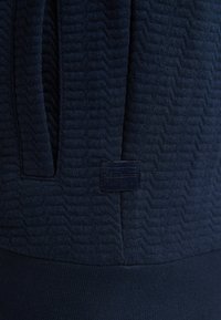 Námořnická modrá mikina s texturovaným vzorem ve tvaru zigzagu, vyrobená z měkké látky a se šitým štítkem s názvem značky na spodní straně.