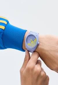 Asmuo reguliuoja lavandinį laikrodį su geltonu Adidas logotipu, vilkėdamas mėlyną striukę su geltonomis juostelėmis ant rankovės.